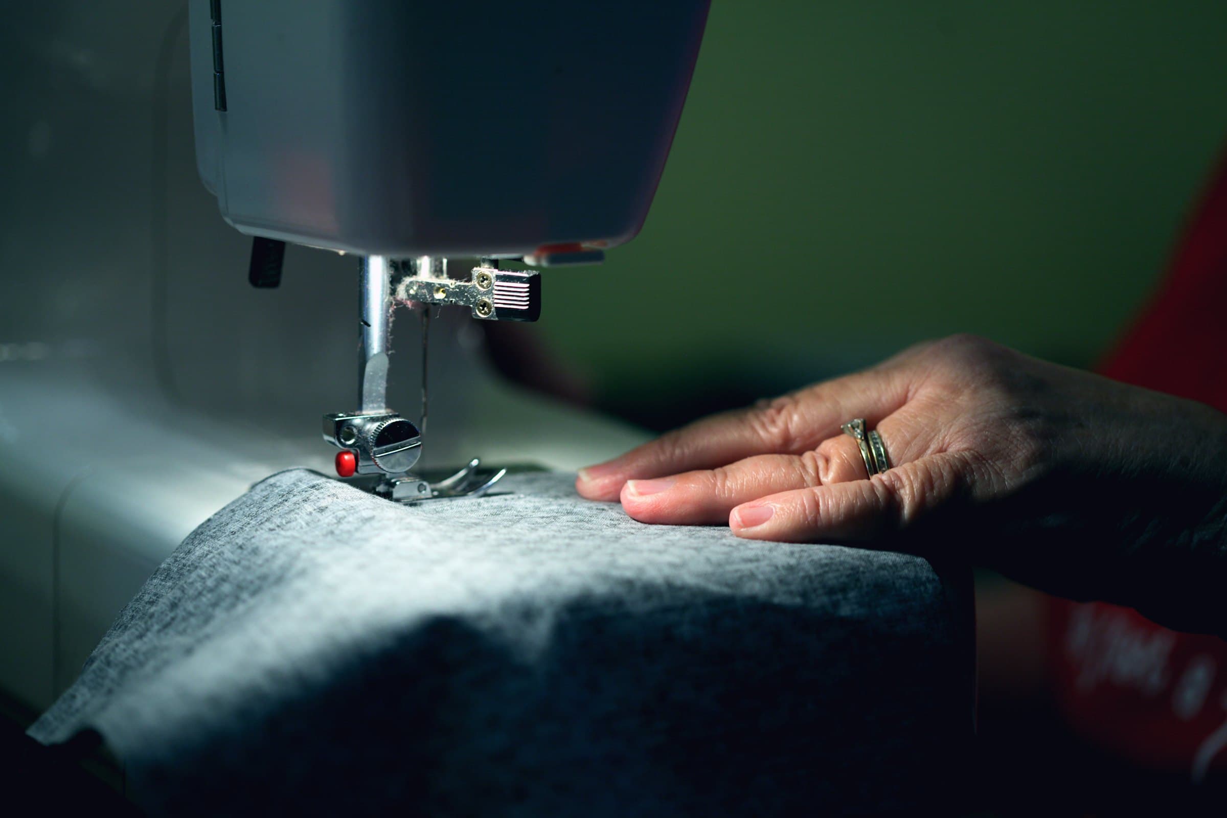 Hands guiding gray felted fabric under a sewing machine needle, lit by the machine lamp against a dark green background