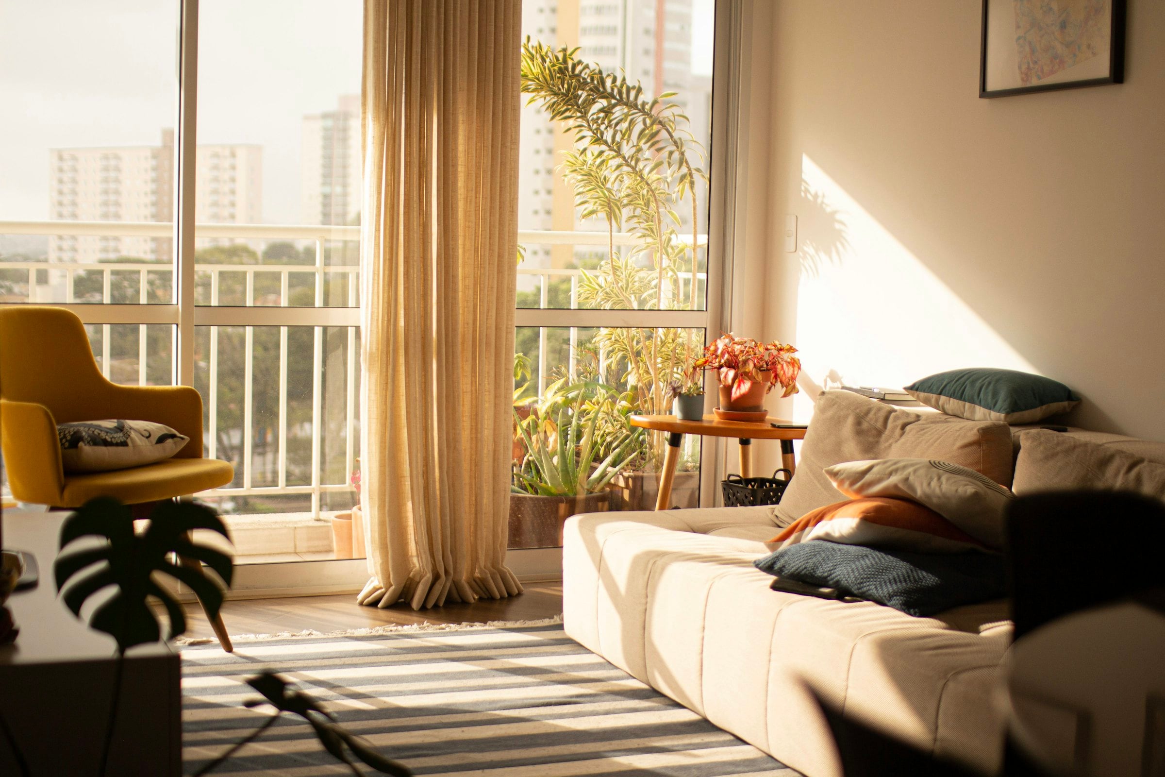 Sunlit living room with floor-length natural linen curtains at a balcony window, a low cream sofa with dark cushions and a mustard armchair, hard afternoon light striping the floor