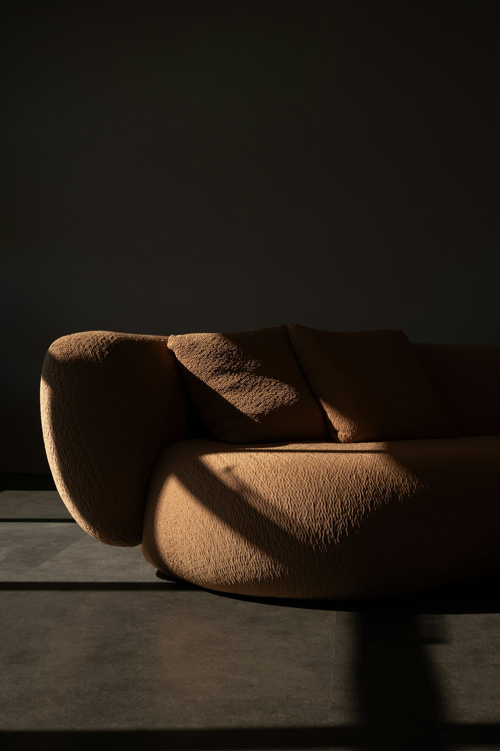 Curved caramel boucle sofa in a dark empty room, cut by a single shaft of raking sunlight