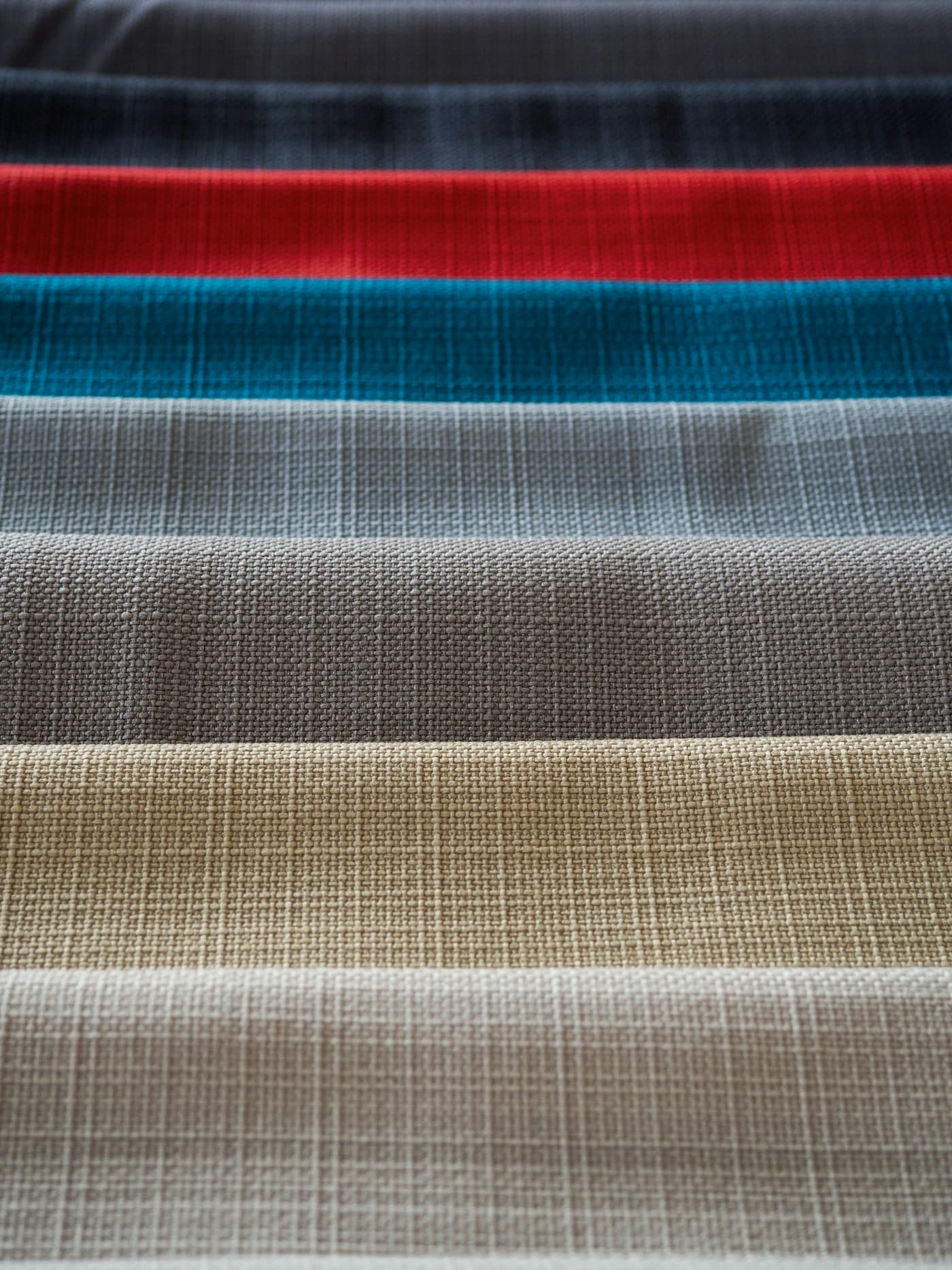 Stacked bolts of textured woven upholstery fabric in bands of charcoal, red, teal, gray, taupe and stone
