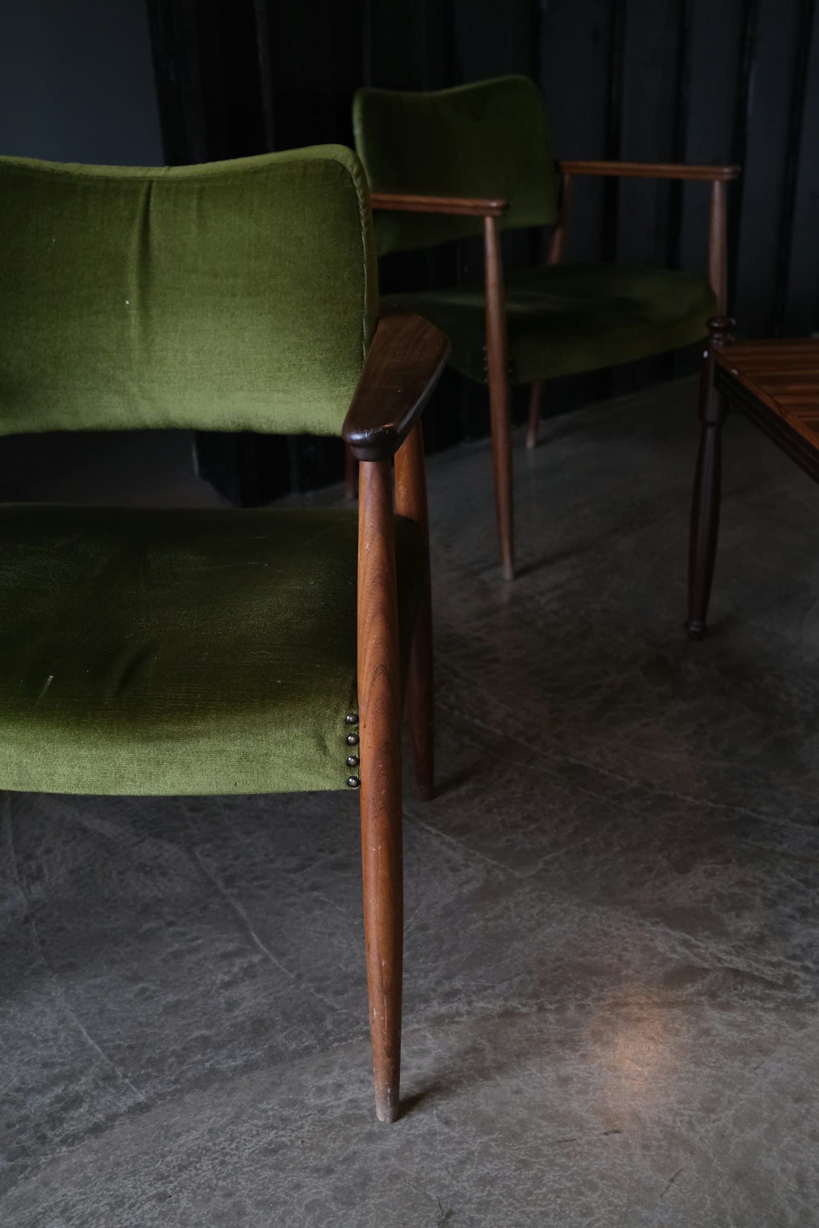 Pair of mid-century walnut armchairs upholstered in moss-green velvet on a polished concrete floor against dark drapery