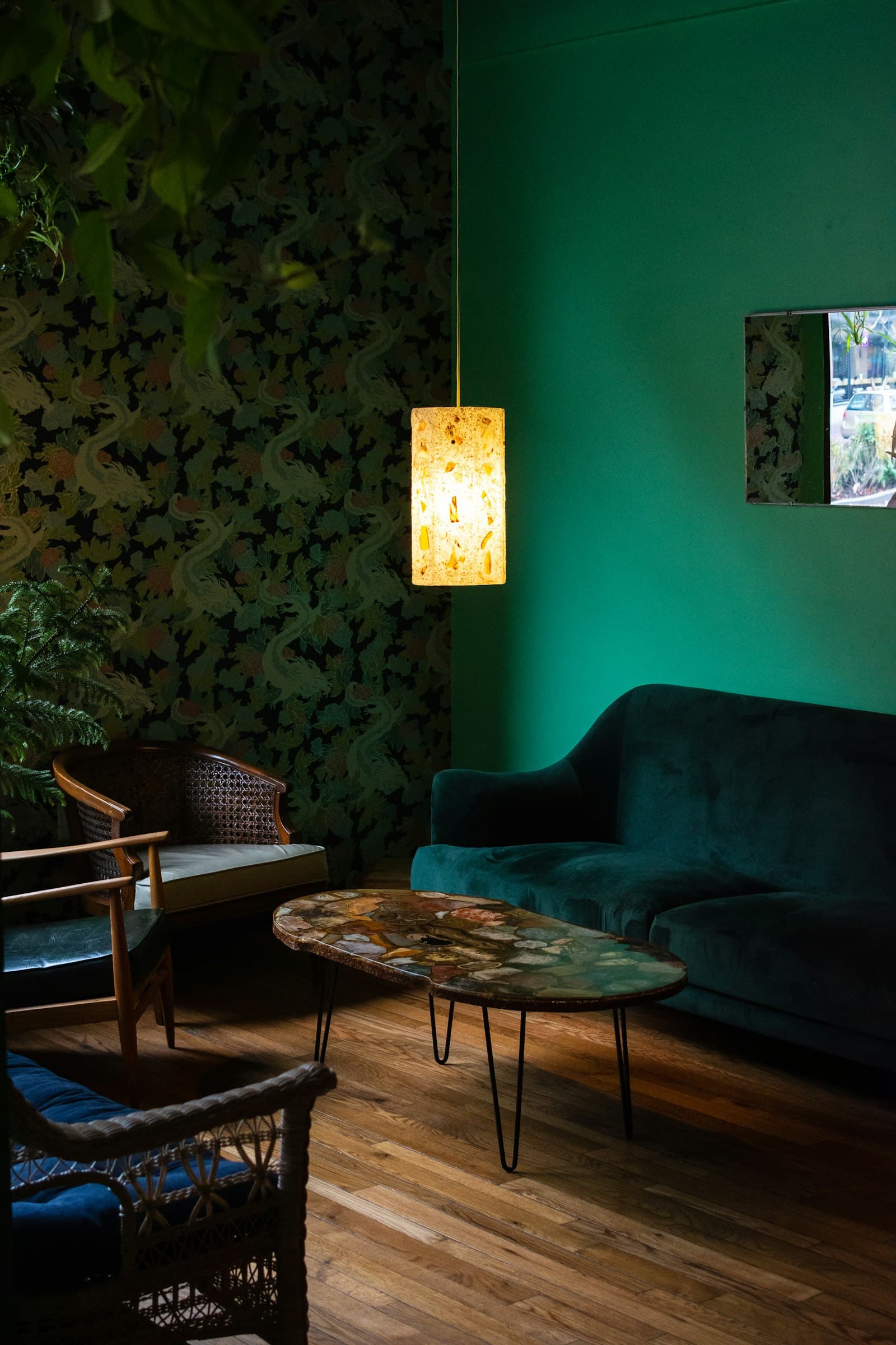 Emerald-green room with a dark green velvet sofa, cane armchairs, an agate-top coffee table, botanical wallpaper and a glowing paper pendant lamp