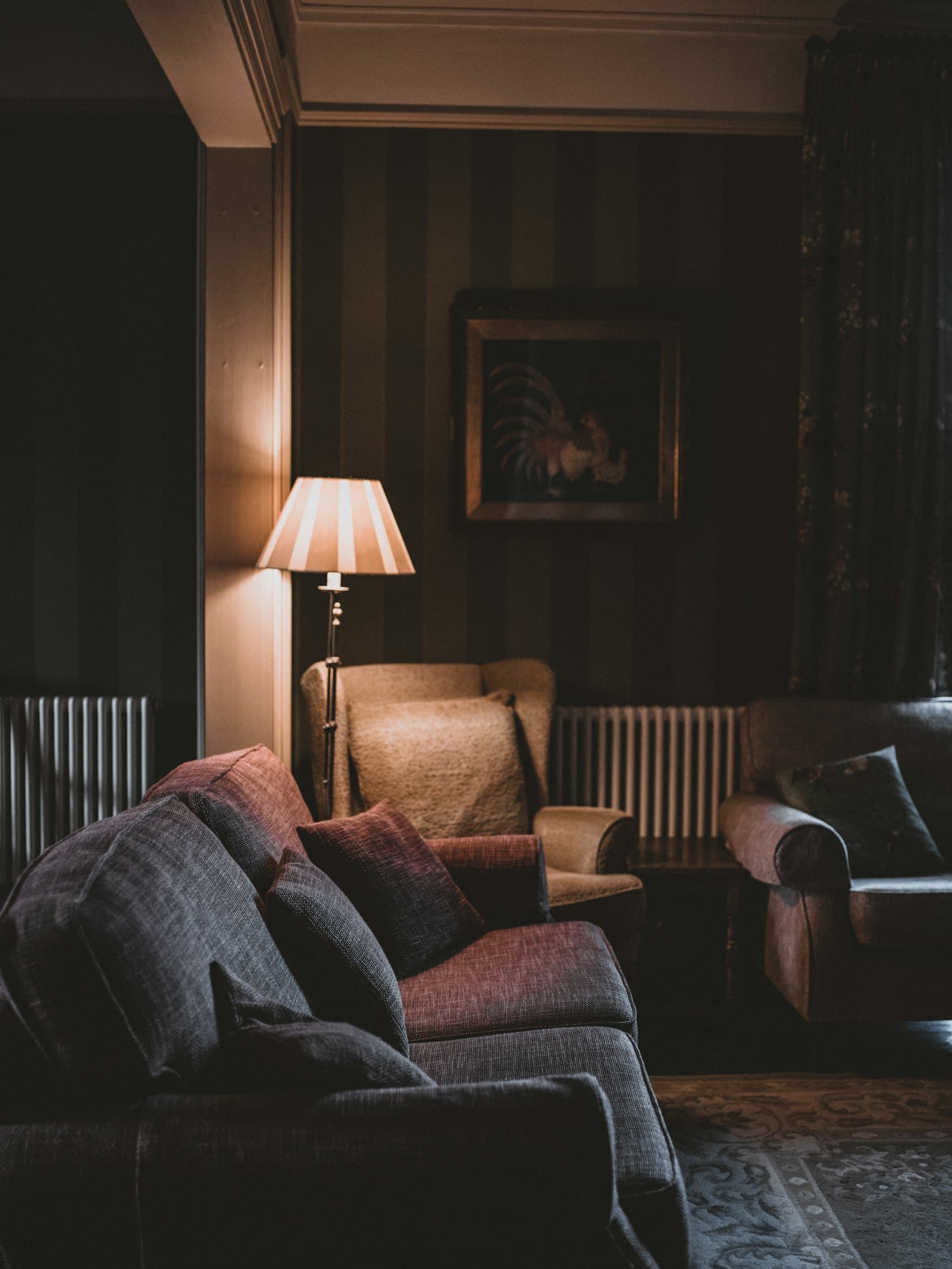 Dim lounge with dark striped wallpaper and heavy curtains, a soft gray sofa with plum cushions and a leather armchair lit by a single pleated-shade floor lamp