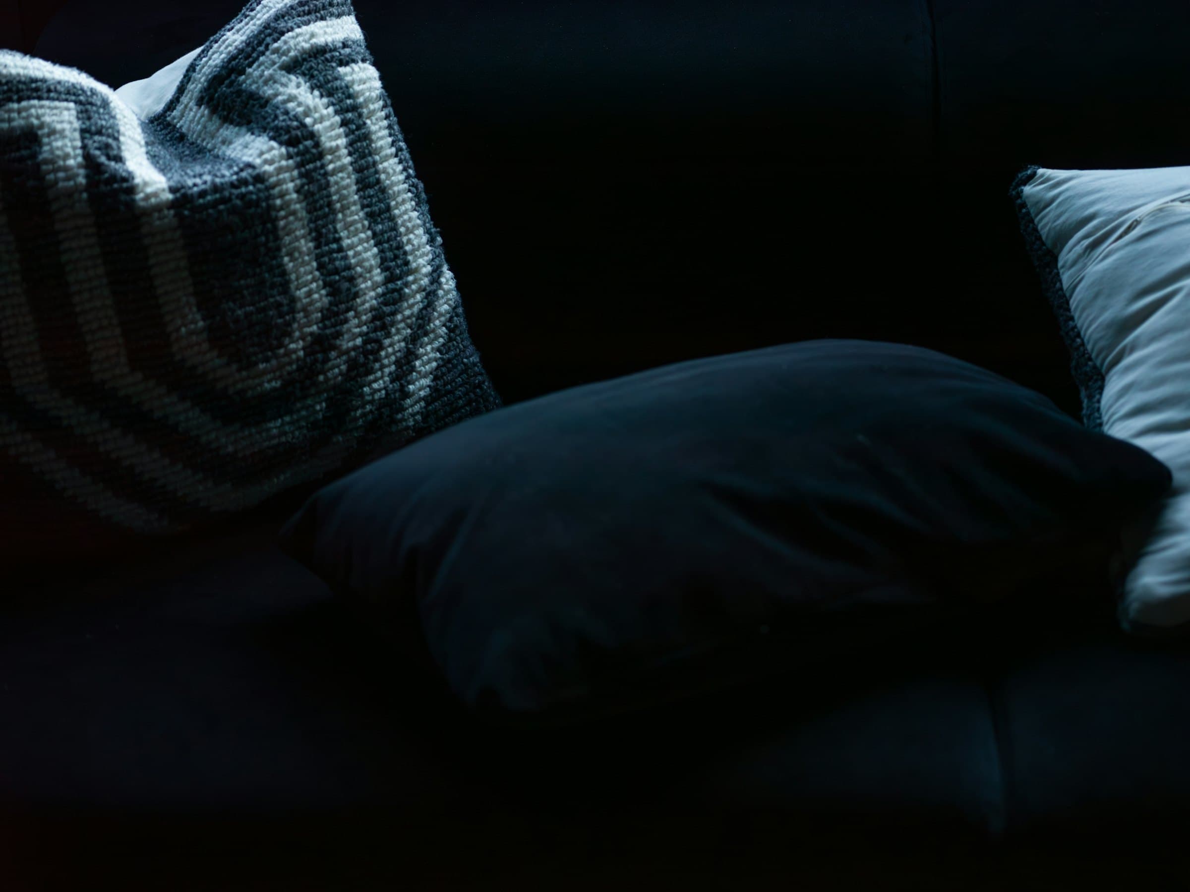 Low-key close-up of a deep navy velvet sofa with a navy cushion and a patterned knit pillow emerging from black shadow