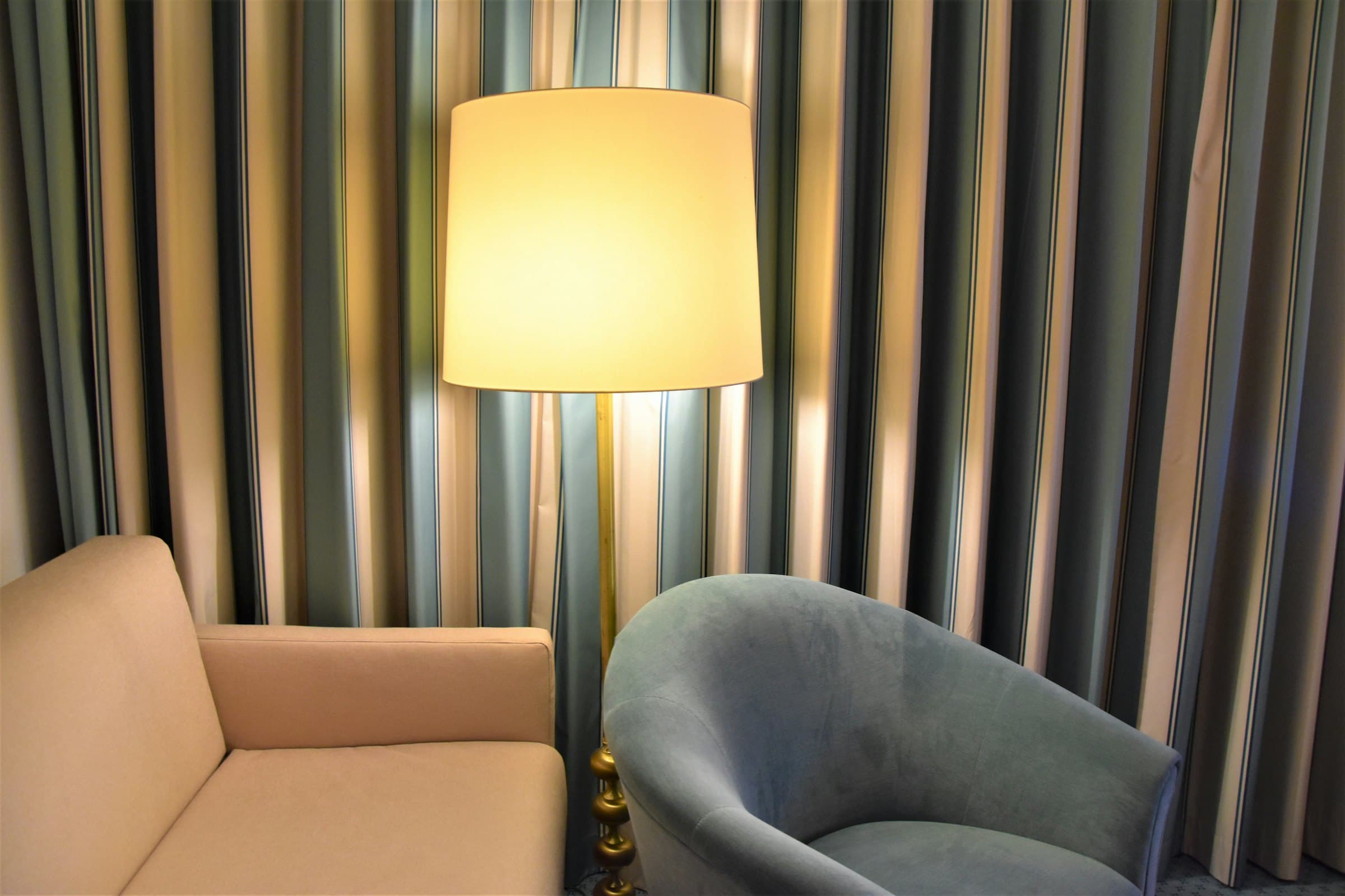 Glowing floor lamp between a blush velvet armchair and a dove-gray velvet tub chair, backed by full-height striped gray and taupe curtains