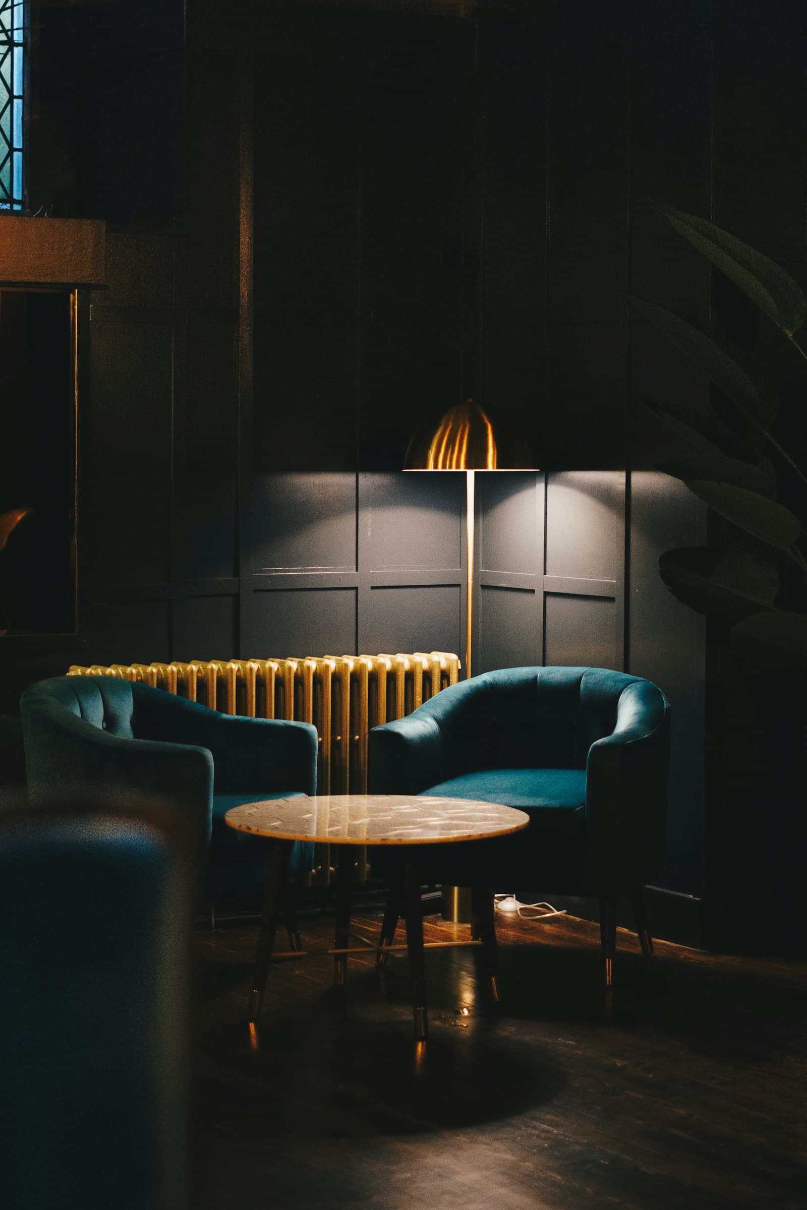 Two teal velvet tub chairs at a round marble table in a near-black panelled room, lit by a brass floor lamp beside a gilded radiator